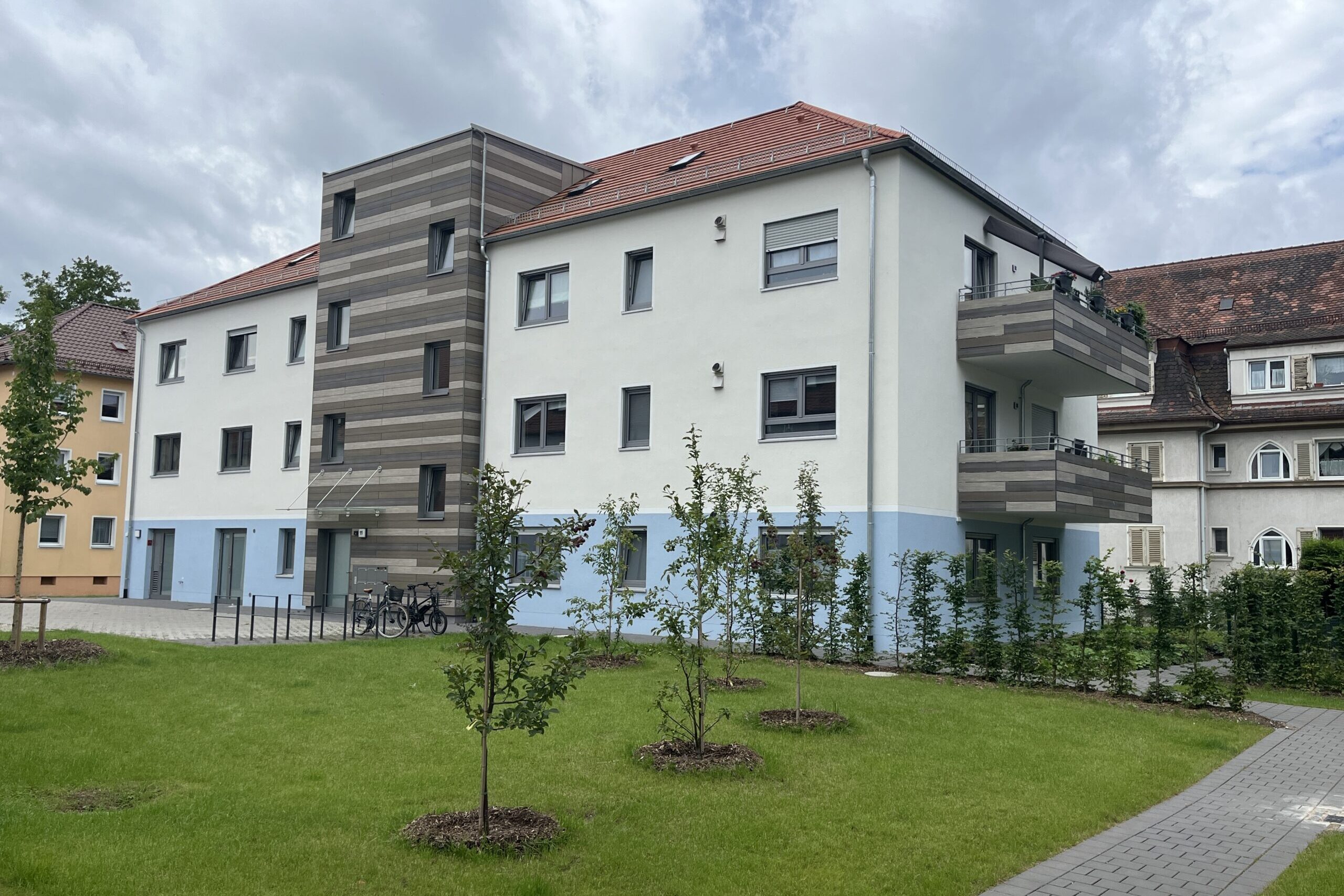 Modernisiertes Mehrfamilienhaus mit Holzfassade und Balkonen, Vorgarten mit neu gepflanzten Bäumen