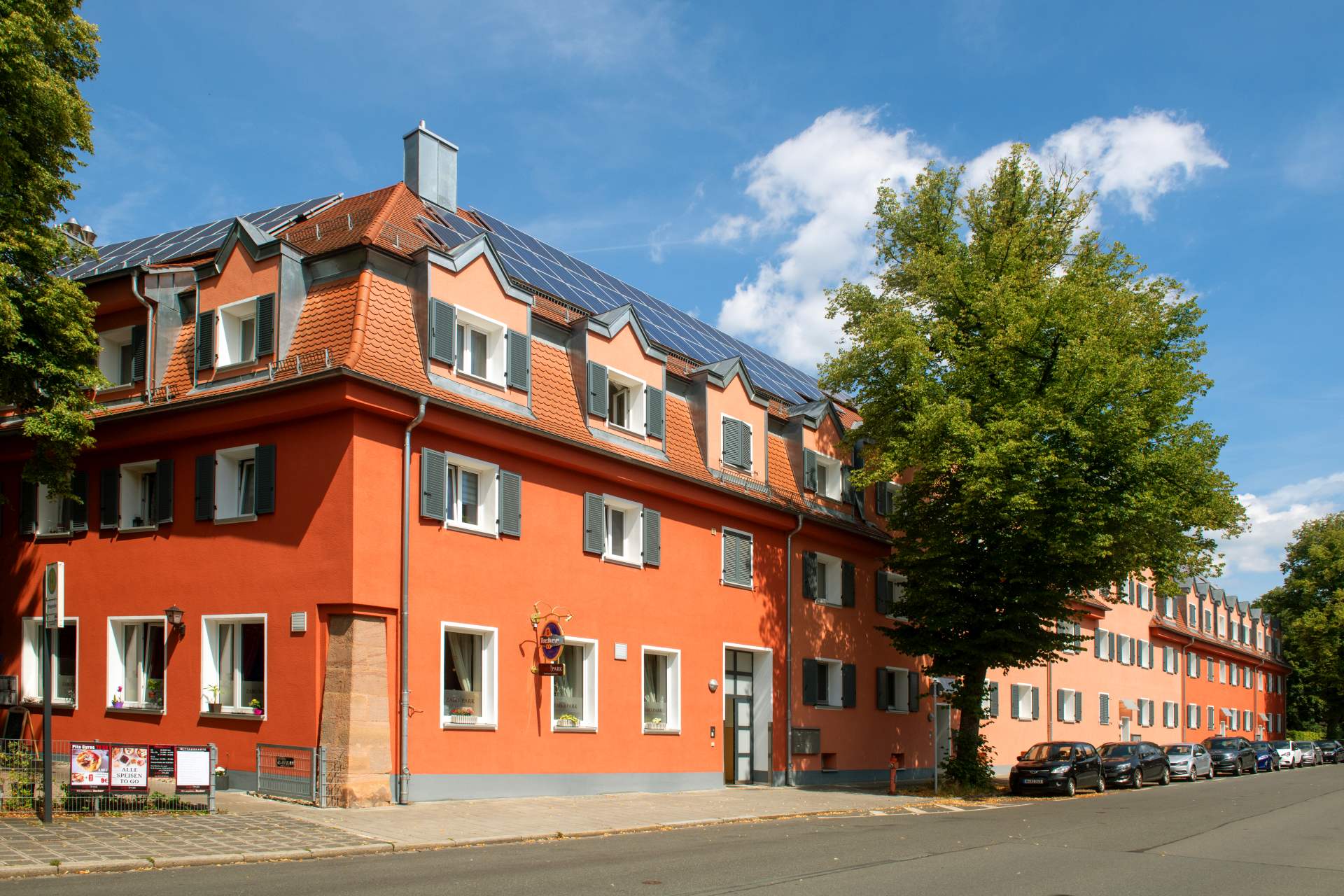 Historisches Gebäude mit orange-roter Fassade, Solaranlage auf dem Dach und Dachgauben, Straßenansicht mit parkenden Autos