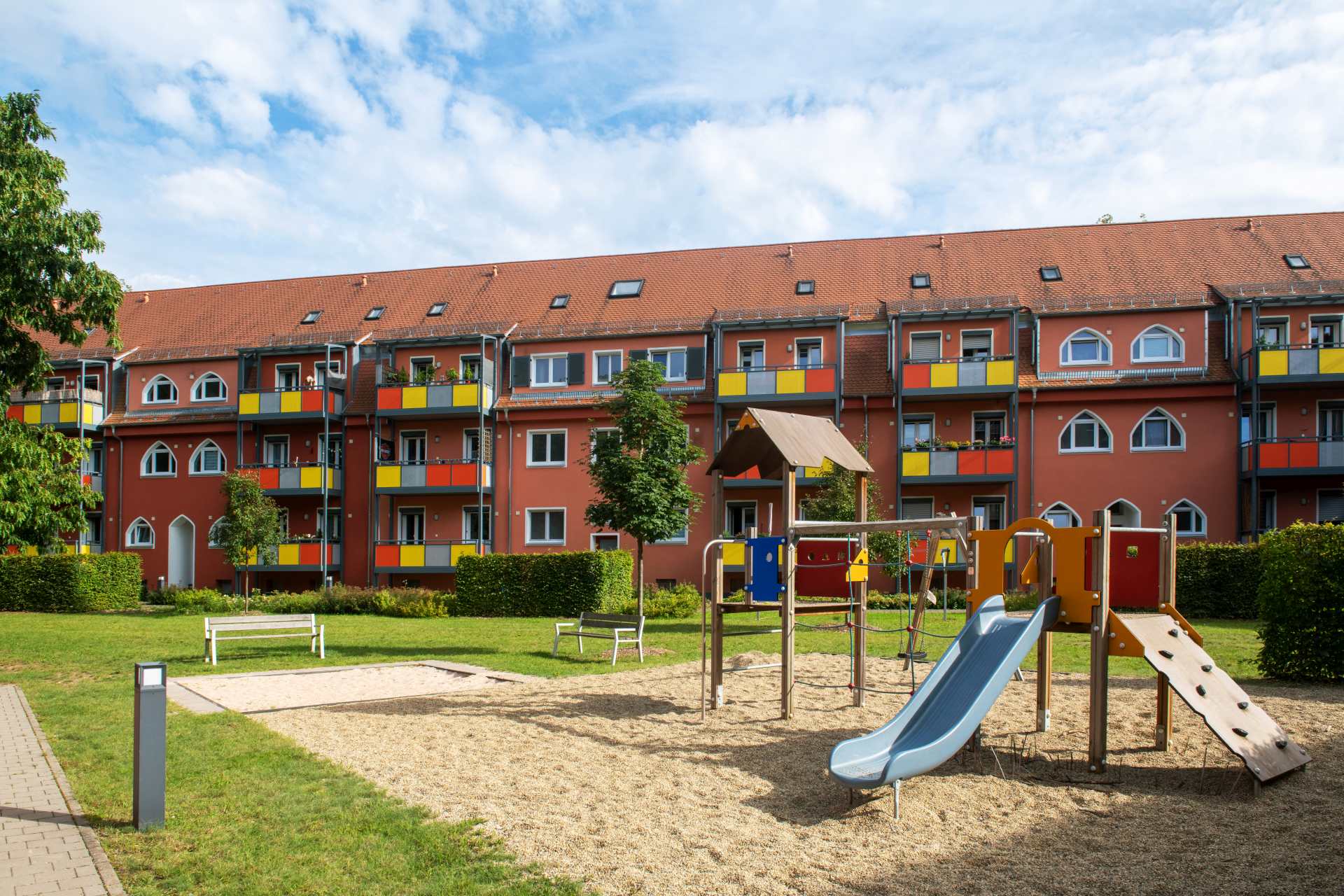 Spielplatz mit Rutsche, Kletterwand und Sitzbänken vor einem roten Wohngebäude mit bunten Balkonen