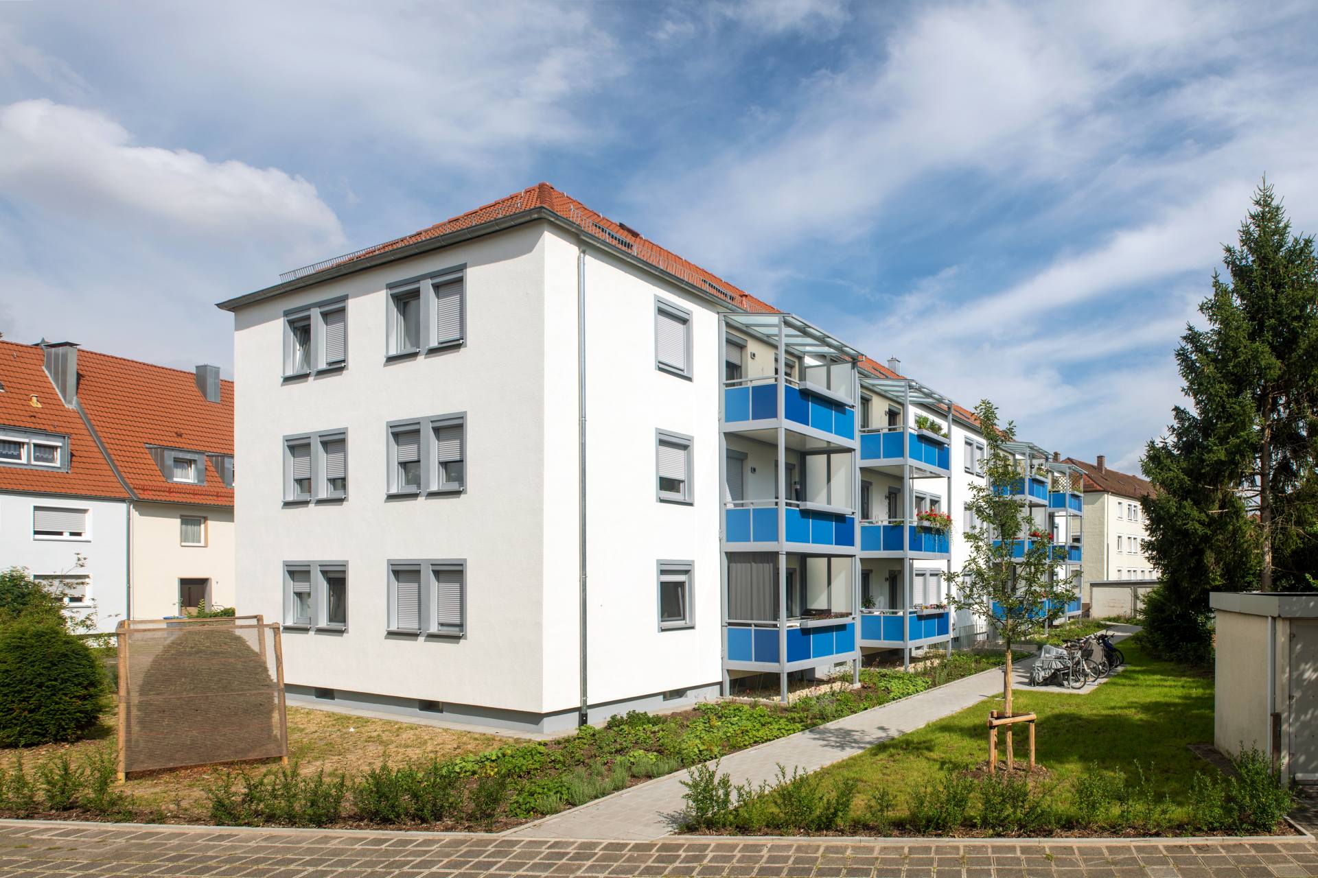 Wohngebäude mit blauen Balkonen und Satteldach, gepflasterter Weg und Grasfläche mit jungen Bäumen im Vordergrund