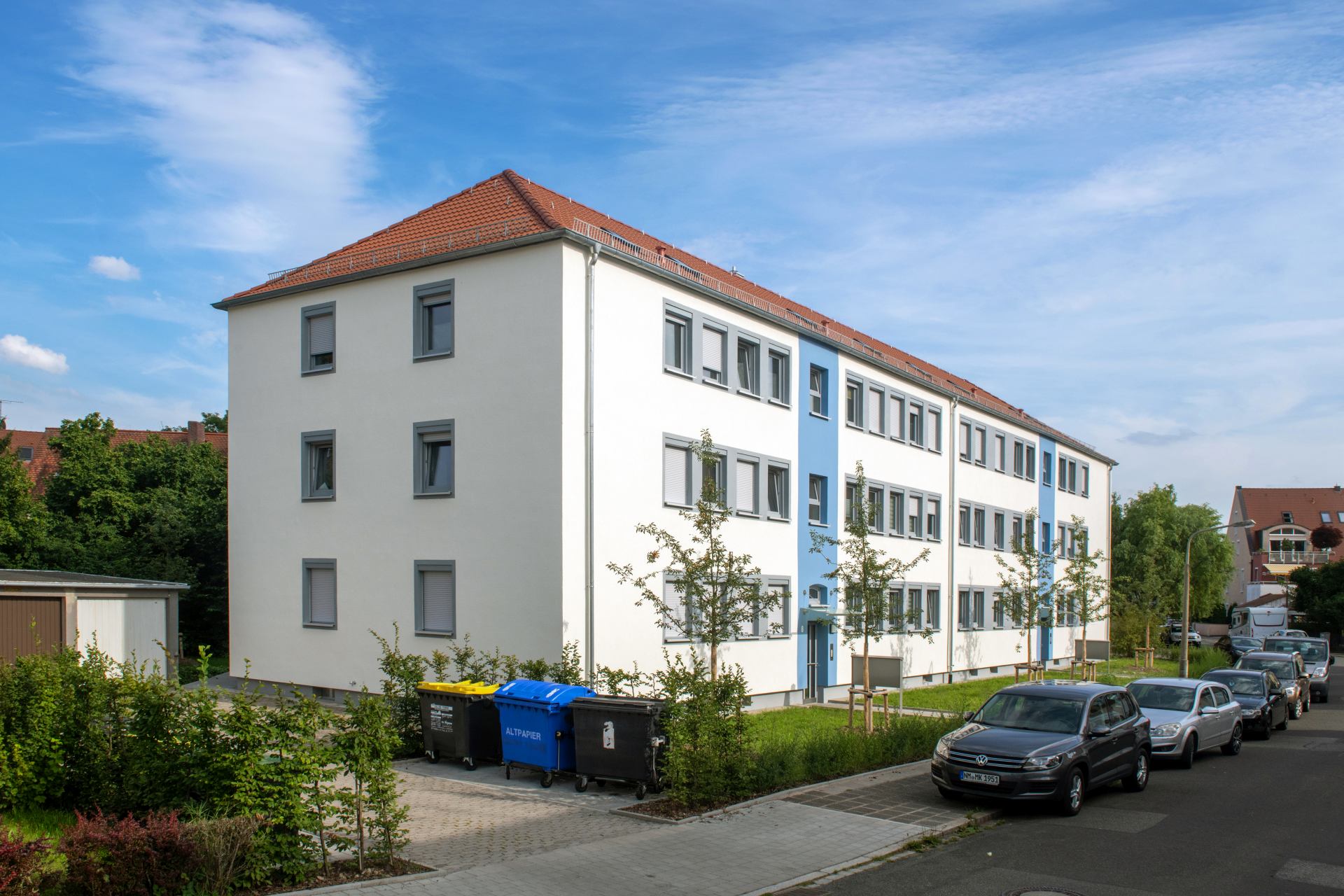 Weißes Wohngebäude mit blauen Fassadenstreifen, Straßenansicht mit Mülltonnen und parkenden Autos