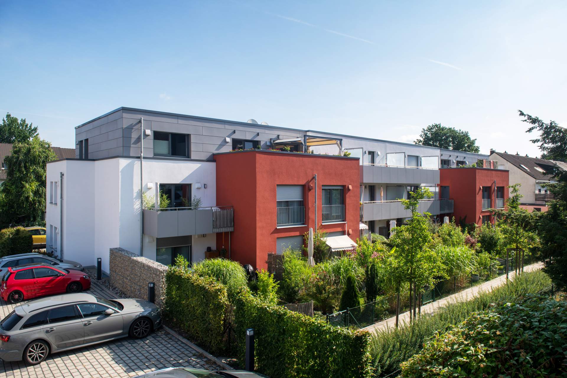 Erhöhte Ansicht des Wohngebäudes mit roter Fassade und Balkonen, dichte Hecken und Parkplatz im Vordergrund