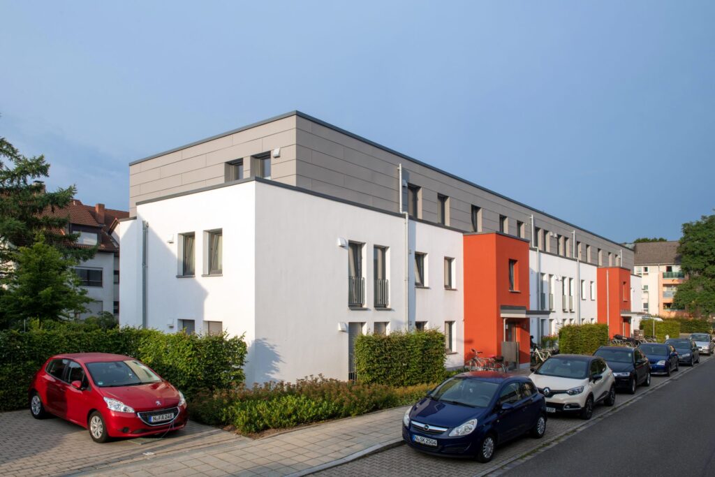 Reihenhauszeile mit weißen, grauen und orangen Fassadenelementen und Flachdach, parkende Autos entlang der Straße