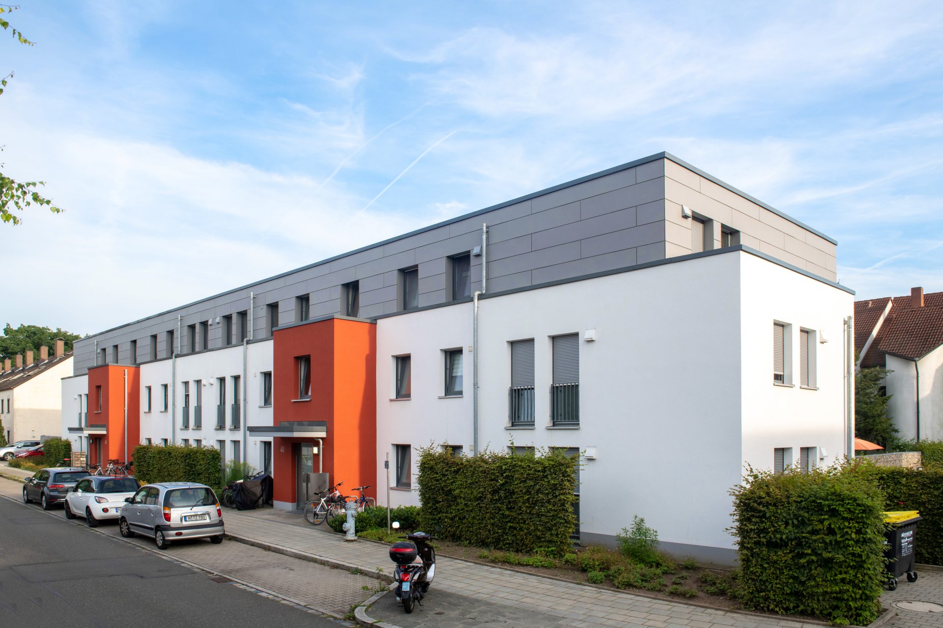 Modernes Wohngebäude mit Flachdach in Weiß, Orange und Grau, davor parkende Autos entlang der Straße