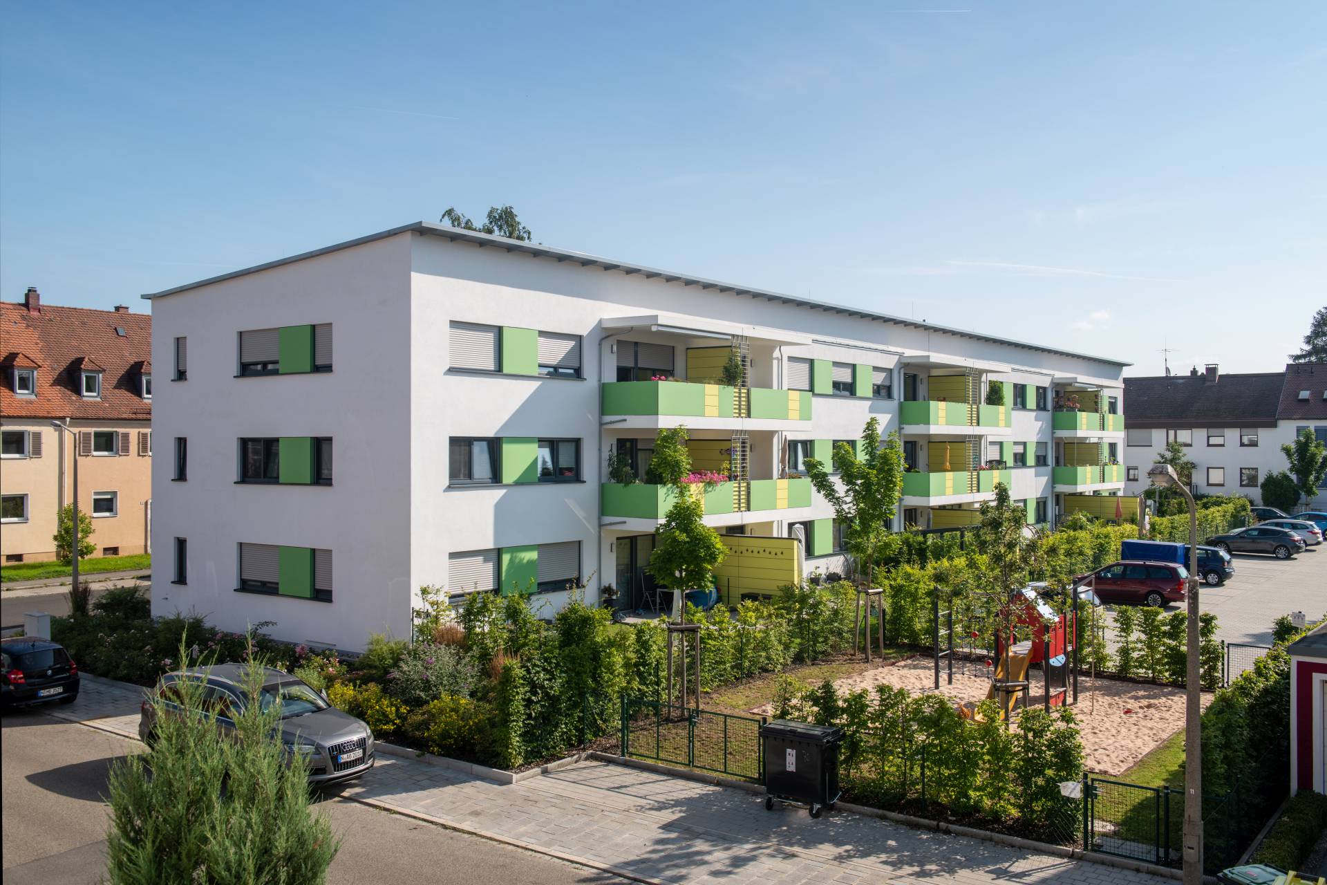 Blick auf das Wohngebäude mit grün-gelben Balkonen, davor ein eingerüzäunter Sandspielplatz und Parkplatz
