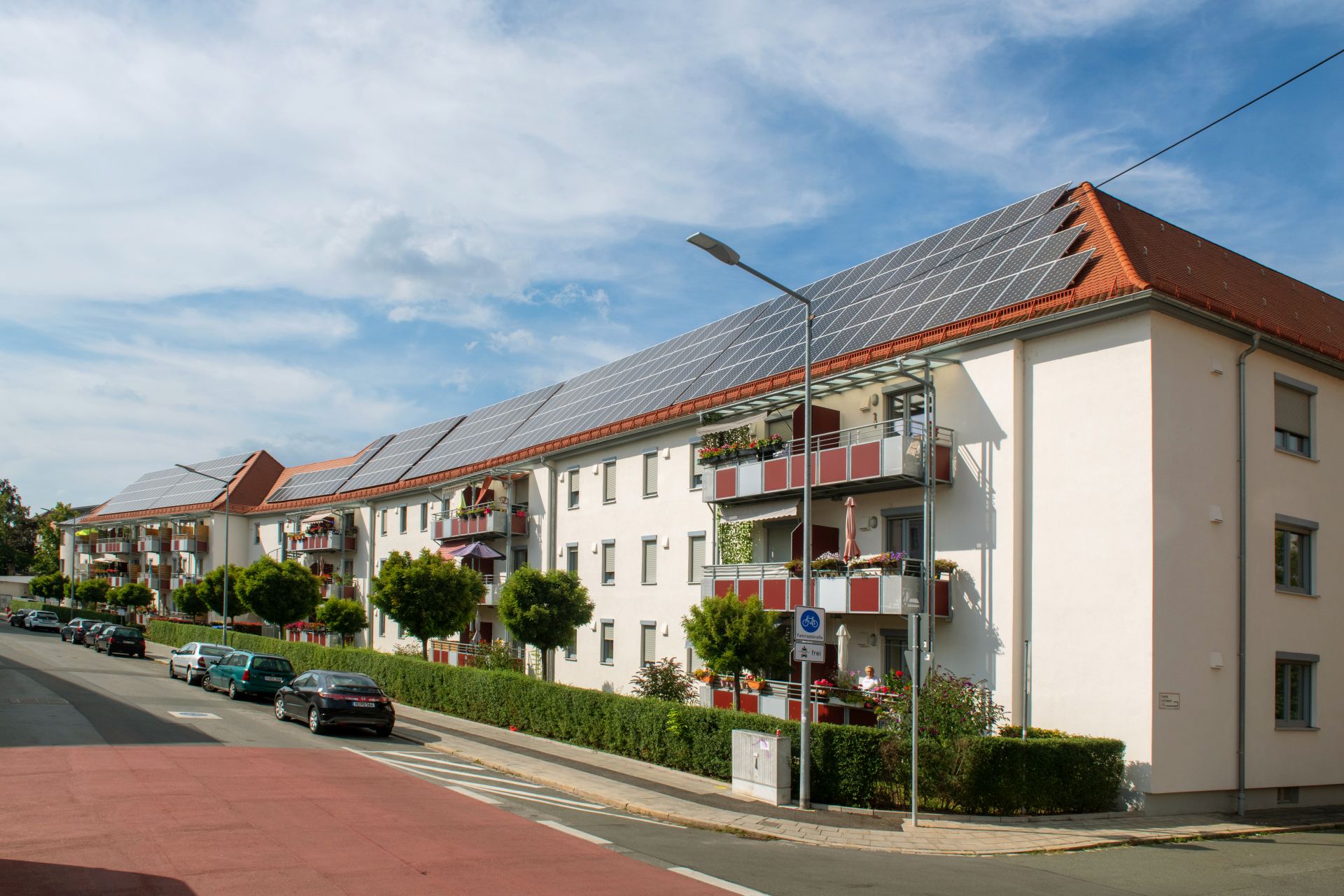 Weißes Wohngebäude mit Solaranlage auf dem Dach und Balkonen, Straßenansicht mit Radweg und parkenden Autos