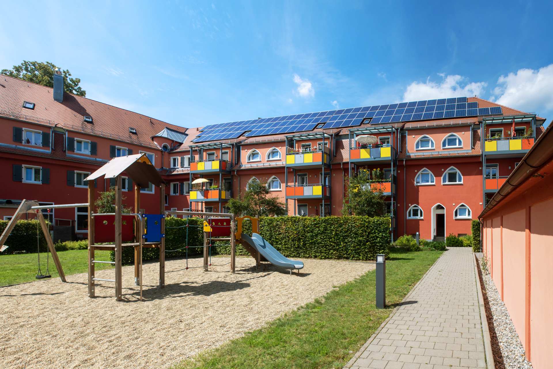 Spielplatz mit Rutsche, Klettergerüst und Schaukel vor einem Wohnhaus mit bunten Balkonen und Solarpaneelen auf dem Dach