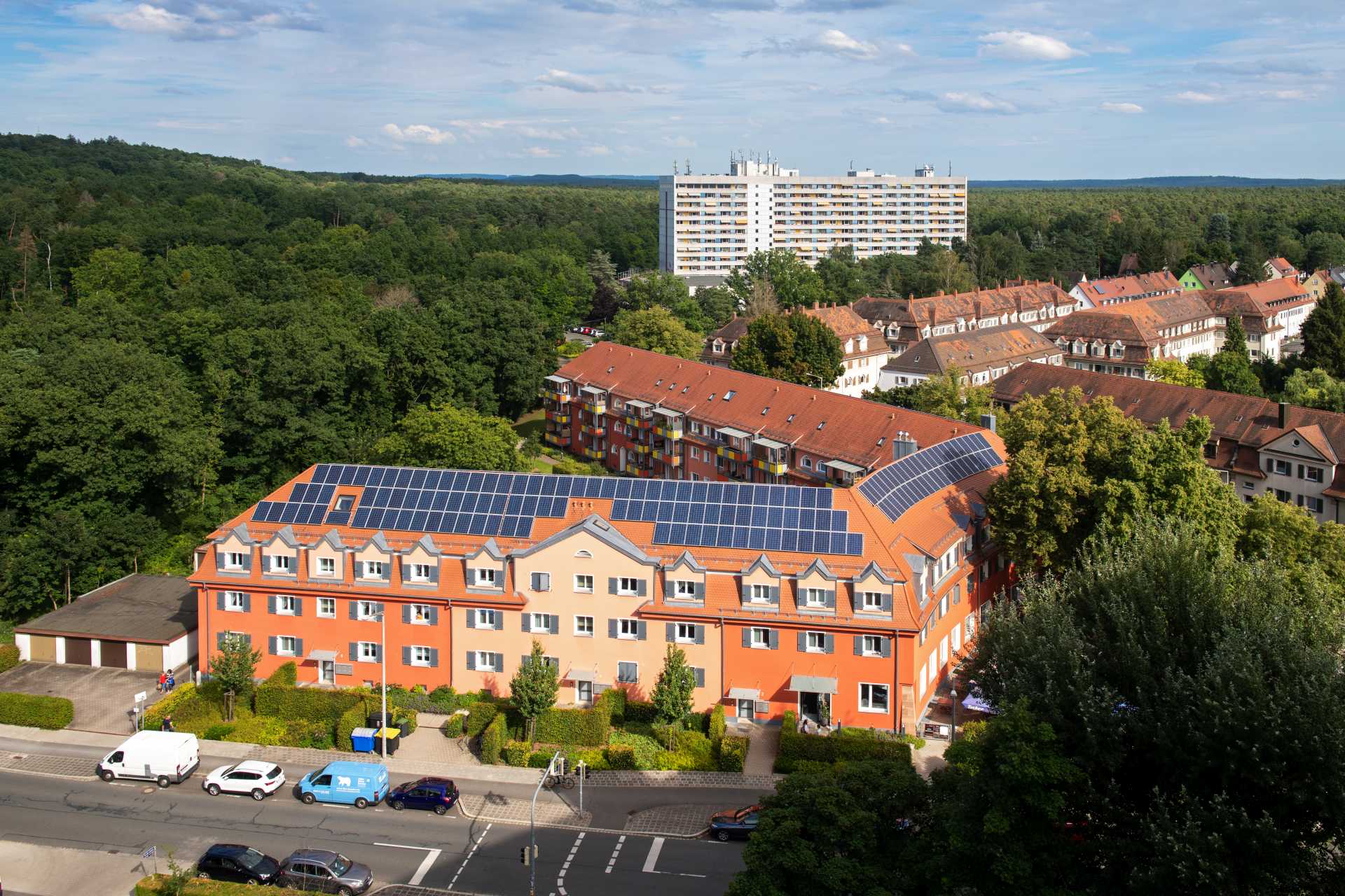 Luftaufnahme der Wohnanlage mit Solarpaneelen auf dem Dach, umgeben von Waldfläche, im Hintergrund ein Hochhaus