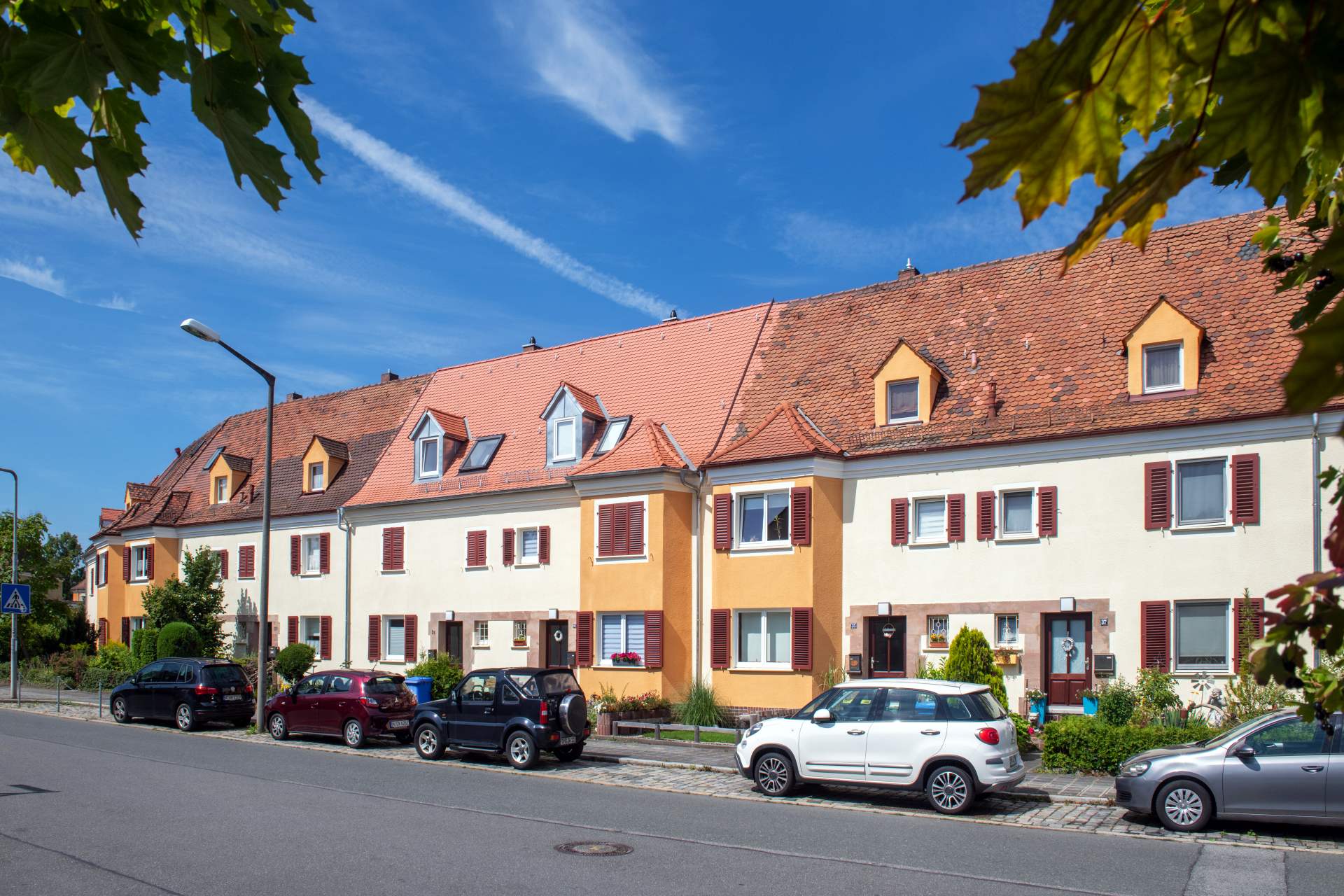 Straßenansicht einer Reihenhauszeile in Gelb- und Ockertönen mit Dachgauben, davor parkende Autos entlang der Straße