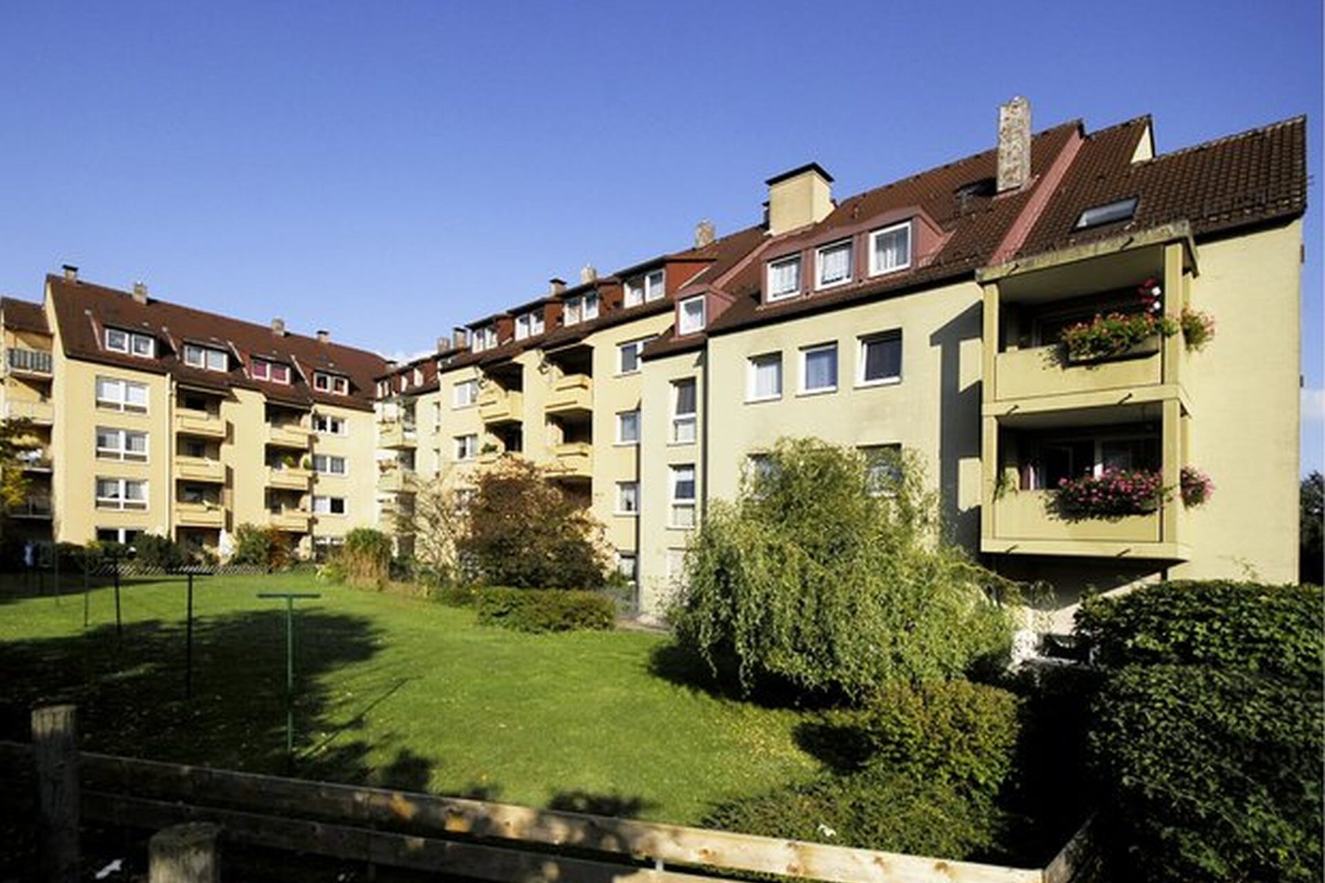 Person sitzt auf einem Sofa und beGelbes Wohngebäude mit Balkonen, Rückansicht mit großer Rasenflächedient ein Tablet mit der Wohnungsunternehmen-Website, unten links das WU-Logo eingeblendet