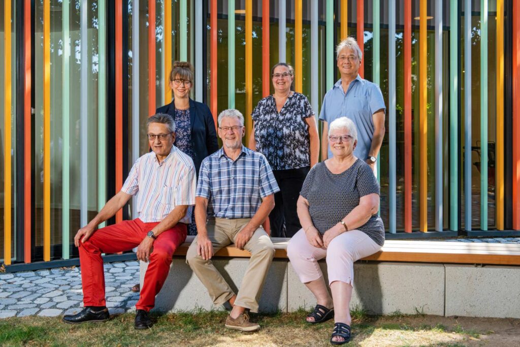 Gruppenfoto von sechs Personen auf einer Bank vor einer bunt gestreiften Fassade